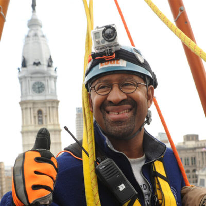 The image shows a man wearing a helmet with a camera attached, safety gear, and glasses. He is smiling and giving a thumbs-up. In the background, there is a tall building with a clock tower. The man appears to be engaged in an outdoor activity, possibly a construction or maintenance task.